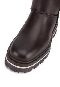 Tmavo hnedá kožená obuv s masívnou zvrškovou podrážkou a bielym prúžkom na boku, s okrúhlou štvorcovou špičkou a viditeľným šitím pri členku.