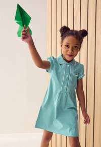 En pige iført en grøn gingham-kjole med krave og knapper, der holder en foldet grøn papirflyver. Baggrunden er af træpaneler.