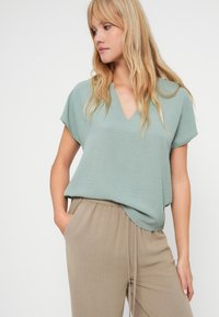 Bluză texturată de culoare verde deschis cu decolteu în V și mâneci scurte, asociată cu pantaloni bej cu talie elastică. Design simplu, croi lejer.