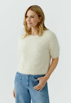 Femme aux cheveux blonds mi-longs portant un pull court à manches courtes blanc en tissu duveteux et un jean bleu, avec une main dans la poche, regardant légèrement vers la gauche.