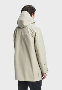 Svijetlosmeđa vodootporna jakna s kapuljačom, koja ima glatku teksturu, minimalistički dizajn i logo naglašene na rukavu i donjem rubu.