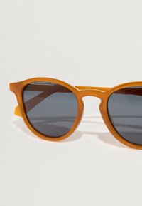 Στρογγυλά γυαλιά ηλίου με πορτοκαλί πλαστικούς σκελετούς, σκούρους γκρι φακούς και με μια γέφυρα που συνδέει τους φακούς. Ελαφριά, με λεία υφή.