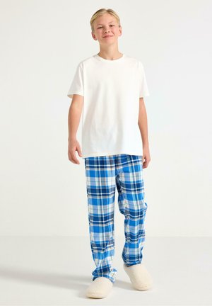Vaaleahiuksinen poika seisoo pukeutuneena yksiväriseen valkoiseen t-paitaan, sinivalkoruudullisiin pyjamahousuihin ja valkoisiin pörröisiin tohveleihin yksinkertaista taustaa vasten.