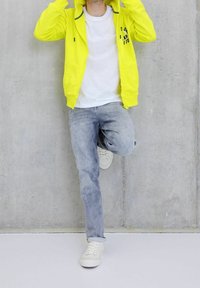 Felpa gialla brillante con zip, t-shirt bianca, jeans grigio chiaro e sneakers bianche. La felpa presenta una stampa sul petto.