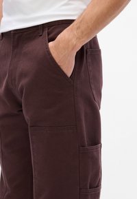 Pantaloni in cotone bordeaux con taglio dritto, tasche laterali e finitura testurizzata. Comprendono due grandi tasche a toppa sulle cosce.