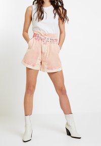 Pantalones cortos de mezclilla rosa claro con una cintura ancha con cinturón, pliegues frontales y detalles en un rosa más oscuro en contraste, combinados con una blusa sin mangas blanca y botines blancos hasta el tobillo.