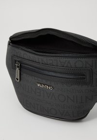 Pochette en tissu noir avec une surface texturée, fermeture zippée sur le dessus et un logo en relief. Dispose d'une poche zippée extérieure pour le rangement.
