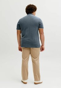 Άνδρας με σγουρά μαλλιά που στέκεται με την πλάτη, φοράει γκρι κοντομάνικο πόλο, μπεζ παντελόνι και λευκά αθλητικά παπούτσια σε απλό φόντο.