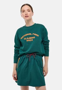 Sweat-shirt vert avec "ZONE D'ESCALADE DU PARC NATIONAL" en texte orange. Assorti à une jupe bleu-vert avec un cordon de serrage et des poches latérales zippées.