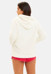 Hoodie texturé blanc avec capuche, présentant un motif en diamant. Le modèle le porte avec un short rose, montrant un design à la fois confortable et polyvalent.