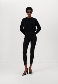 Suéter negro de punto, leggings negros de talle alto y zapatos de tacón alto negros con acentos plateados. Las gafas de sol completan el conjunto.