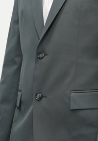 Σκούρο γκρι σακάκι κοστουμιού με διακριτή υφή, διακοσμημένο με δυο μαύρους κουμπιά, με κλασικές πέτε�ς και μια μόνο τσέπη στο στήθος.