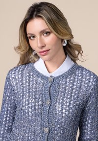 Cárdigan de punto en azul claro con un intrincado diseño de calados, detalles en botones plateados y un cuello redondo sobre una camisa blanca.