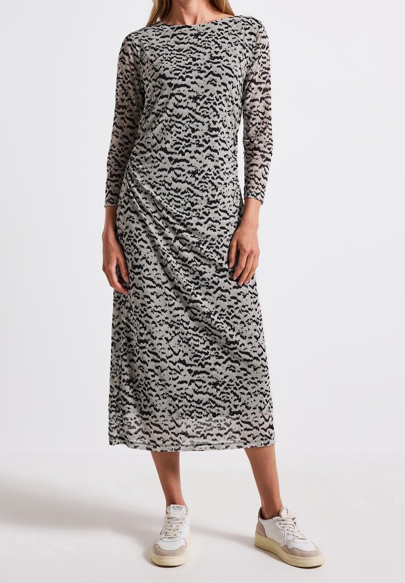 Femme portant une robe grise mi-longue avec un imprimé abstrait noir et des manches longues, associée à des baskets blanches et beiges.