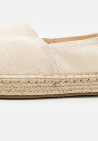 Espadrille de lona beige con suela de yute trenzado, presenta un upper texturizado y detalles cosidos a lo largo del borde de la suela.