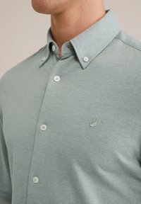 Světle tyrkysová košile na knoflíky z texturované látky s špičatým límečkem, bílými knoflíky a malým vyšívaným logem na hrudi.