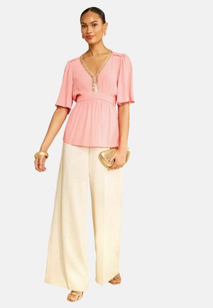 Donna con camicetta rosa a scollo a V con bordatura dorata, pantaloni crema a gamba larga, sandali dorati, braccialetto e pochette, in piedi su sfondo bianco.