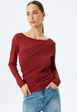 Top bordeaux a maniche lunghe con design drappeggiato e dettagli arricciati in vita, abbinato a jeans in denim blu chiaro. Tessuto morbido e vestibilità comoda.