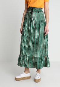 Jupe longue verte à motifs avec un ourlet à volants en plusieurs couches, présentant des dessins géométriques en crème et noir, associée à des baskets blanches à plateforme.