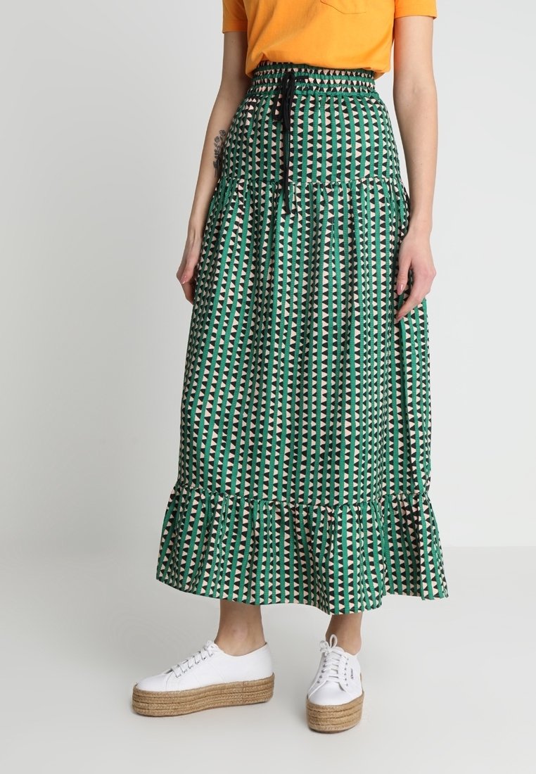 Jupe longue verte à motifs avec un ourlet à volants en plusieurs couches, présentant des dessins géométriques en crème et noir, associée à des baskets blanches à plateforme.