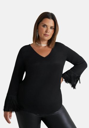 Schwarzes Oberteil mit V-Ausschnitt und langen Ärmeln, verziert mit schwarzen Fransenakzenten an den Bündchen. Hergestellt aus glattem Stoff, kombiniert mit Leggings aus Kunstleder.