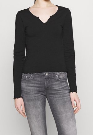 Femme portant un haut noir ajusté à manches longues avec col en V, associé à un jean gris délavé taille haute, debout devant un fond uni.