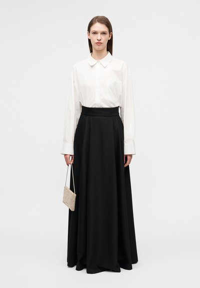 Άσπρο πουκάμισο με κουμπιά και μακριά μανίκια, συνδυασμένο με μαύρη maxi φούστα με ψηλή μέση. Το μοντέλο κρατά μια μικρή, ανοιχτόχρωμη, υφασμάτινη τσάντα.