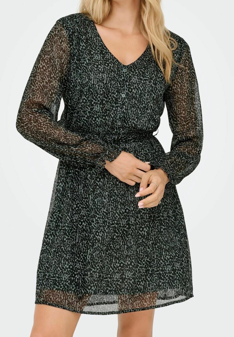 Femme portant une robe longue à manches longues transparente noire et grise à motifs, avec des boutons et une taille cintrée, debout devant un fond uni.