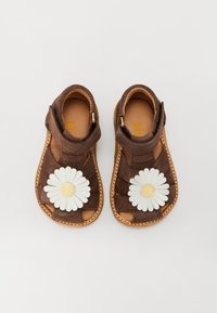 Sandales en daim marron avec un design tissé, ornées d'une grande marguerite blanche avec un centre jaune sur chaque chaussure et de semelles beige. Fermeture par bande Velcro.