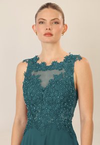 Vestido de noche en color teal con bordado de encaje floral y acentos de cuentas, un escote transparente y una silueta ajustada.