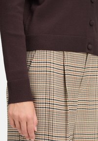 Brown knitted cardigan with ribbed cuffs, worn over beige plaid trousers featuring black and yellow accents and pleats.