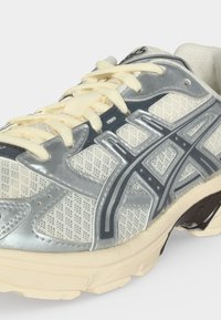 Κοντινή λήψη ενός λευκού και ασημί αθλητικού παπουτσιού με εκρού κορδόνια και μαύρο λογότυπο Asics στο πλάι και μπεζ σόλα.