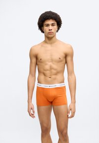 Πορτοκαλί ανδρικά μποξεράκια με άσπρη ζώνη μέσης που φέρει το λογότυπο "TOMMY HILFIGER". Το ύφασμα φαίνεται λείο και ελαστικό. Το μοντέλο στέκεται προς τα εμπρός.