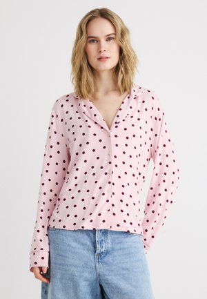 Femeie blondă purtând o bluză roz cu nasturi, cu buline închise la culoare, și blugi din denim albastru deschis, stând în picioare pe un fundal alb.