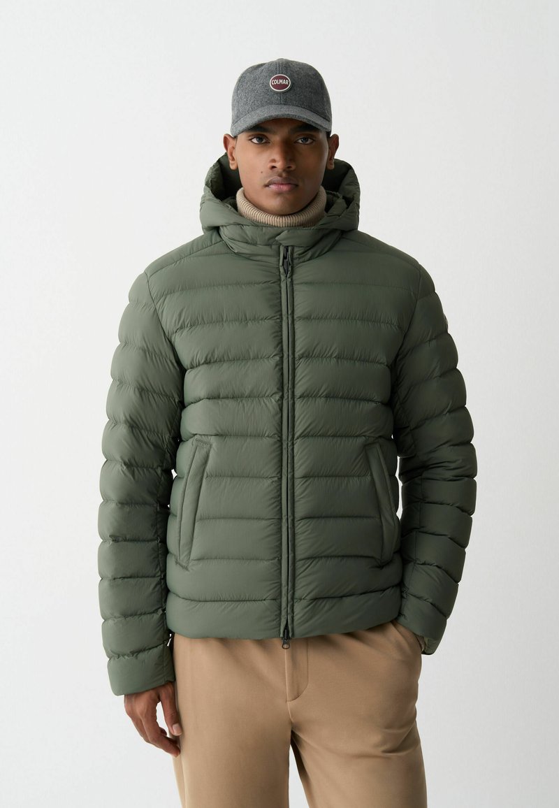 Olíva zöld közepes vastagságú dzseki steppelt mintázattal, elülső cipzárral és kapucnival. Világos bézs garbóval és nadrággal párosítva.