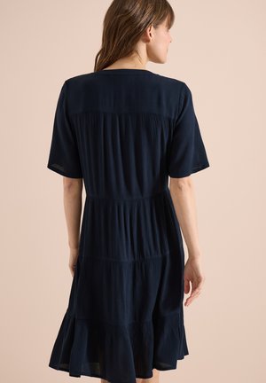 Frau mit braunen Haaren, die ein kurzärmeliges, knielanges, marineblaues Stufenkleid trägt und mit dem Rücken zu einem beigen Hintergrund steht.