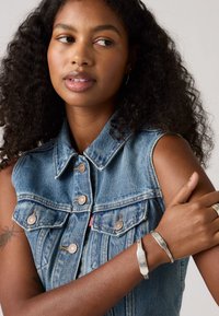 Le gilet en denim présente un délavage bleu clair, des boutons métalliques et un design avec col. Le mannequin porte des bracelets en argent au poignet.