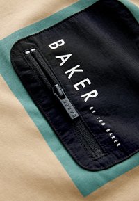 Écusson en tissu noir avec fermeture éclair et texte blanc « BAKER by Ted Baker » sur tissu beige à rayures vertes.