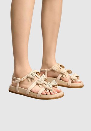 Sandales plates beiges avec brides à la cheville présentant trois nœuds cloutés, portées pieds nus sur un fond uni.