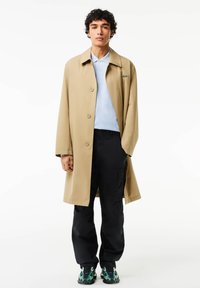 Beige lång kappa, knäppt, med spetsig krage. Ljusblå polo under, svarta cargobyxor och mönstrade gröna-svarta skor.