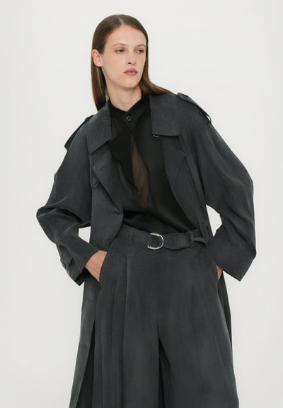 Grijze trenchcoat met een ontspannen pasvorm, schouderen epauletten en een ceintuur in de taille. Draagt over een zwarte doorschijnende top en brede broekspijpen.