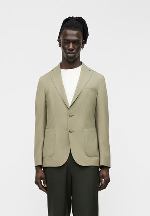 Homme aux cheveux tressés portant un blazer beige sur une chemise blanche et un pantalon foncé, debout devant un fond clair uni.