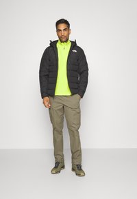 Svart puffjacka över en neongrön dragkedjepolo, tillsammans med beige cargobyxor och bruna vandringsskor. Enkelt design, praktiskt för utomhusbruk.