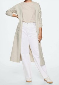 Gilet long beige à ouverture devant, associé à un haut beige clair, un pantalon large blanc et des baskets crème. Tissu doux, coupe décontractée.