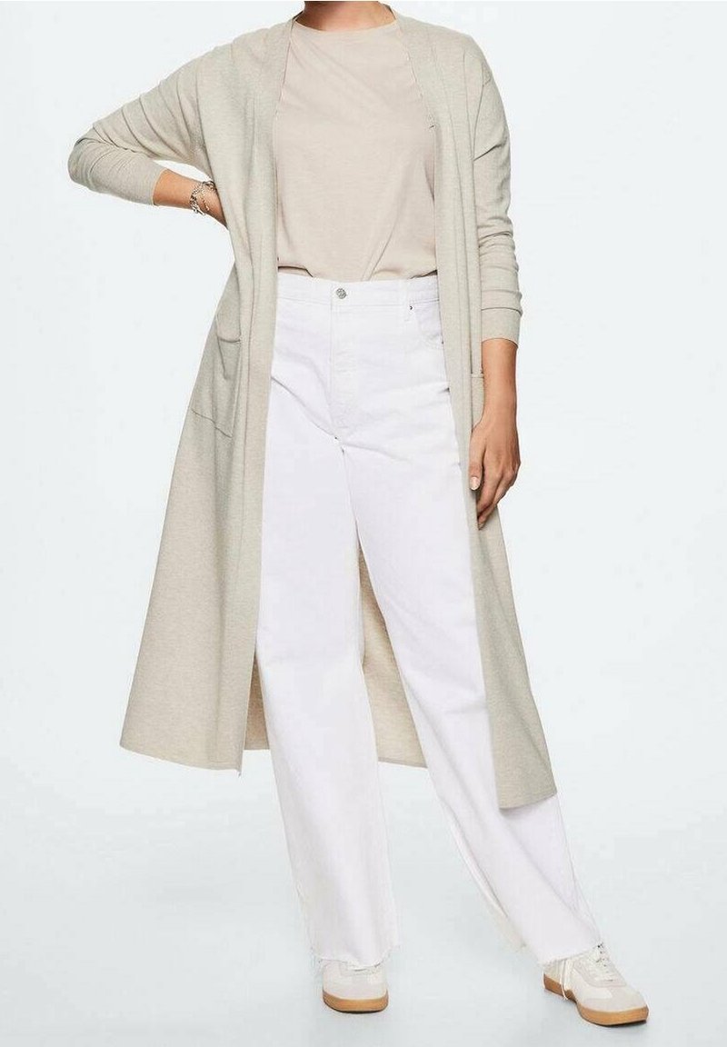 Gilet long beige à ouverture devant, associé à un haut beige clair, un pantalon large blanc et des baskets crème. Tissu doux, coupe décontractée.