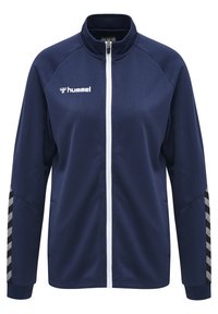 Marinblå zip-up träningsjacka med texturerad tyg, vit dragkedja och kontrasterande svart-vita chevronränder på ärmarna. Hummel-logotyp på bröstet.