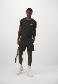 Camiseta deportiva negra con logo blanco, combinada con pantalones cortos negros. El modelo sostiene una raqueta de tenis, vistiendo zapatillas blancas y calcetines hasta el tobillo.