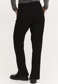 Schwarze Hose mit geradem Schnitt, mit Gesäßtaschen und Gürtelschlaufen, aus einem glatten Stoff gefertigt. Getragen mit schwarzen Schuhen.