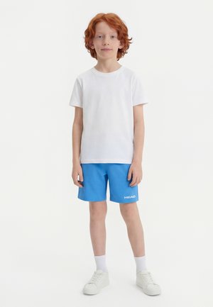 Rdečelasi deček stoji, oblečen v preprosto belo majico, modre kratke hlače z logotipom "HEAD", bele nogavice in bele superge na belem ozadju.