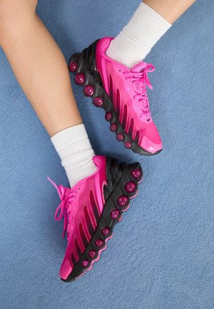 Nogi w białych skarpetkach i jaskrawo różowych butach sportowych z czarnymi podeszwami i amortyzacją przypominającą bąbelki, ustawione na niebieskim dywanie.